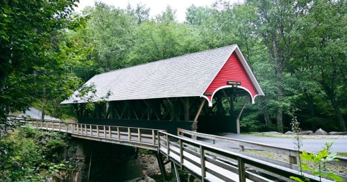 A red covered bridge spans a small creek, surrounded by lush green trees and a wooden railing path.