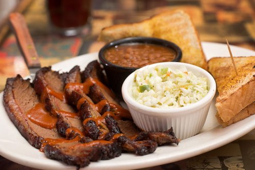 A plate featuring sliced brisket with barbecue sauce, coleslaw, baked beans, and toasted bread.