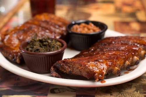 A plate of barbecue ribs with sauce, served with sides of collard greens and baked beans in small bowls.