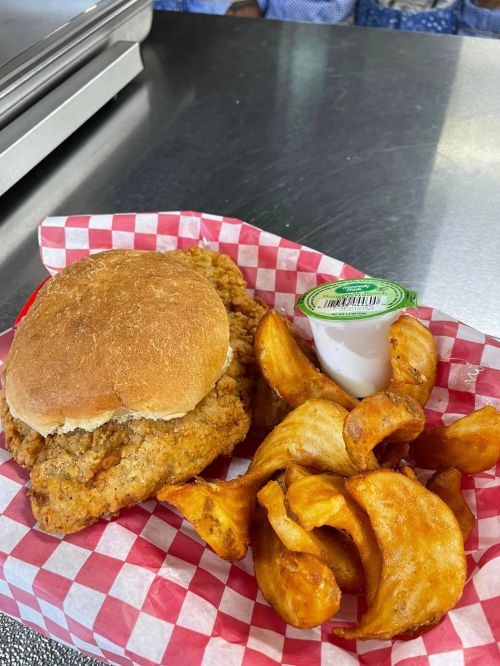 A crispy chicken sandwich on a bun with a side of curly fries and a small container of dipping sauce, served in a red-checkered basket.