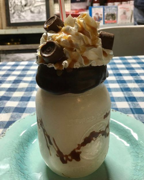 A tall milkshake topped with whipped cream, chocolate, caramel, and candy, served in a mason jar on a blue checkered tablecloth.