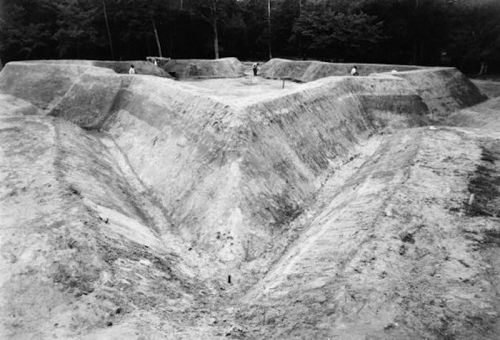 Black and white image of a large, V-shaped excavation site with workers in the background among trees.