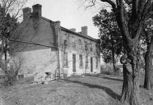 Black and white photo of an old, two-story brick building with a stone foundation, surrounded by trees and grass.