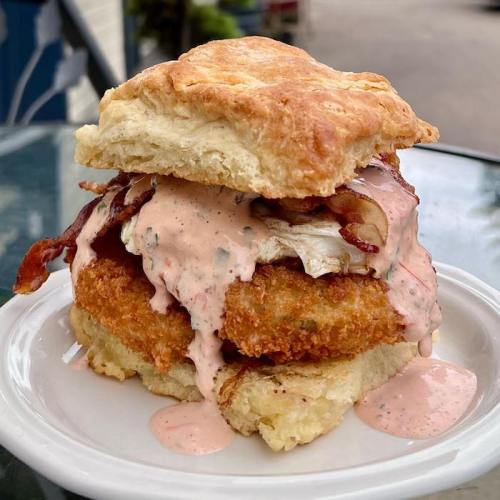 A crispy chicken sandwich with bacon and sauce, served on a flaky biscuit, on a white plate.