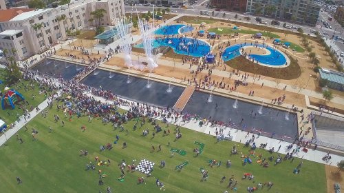 Aerial view of a park with water features, playgrounds, and a large crowd enjoying a sunny day.