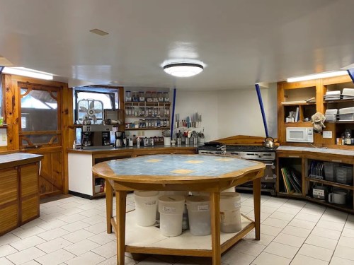 A spacious kitchen with a large round table, shelves of spices, and various appliances, featuring wooden accents.