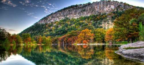 A serene lake reflects a colorful forest and a rocky hill under a clear sky, showcasing autumn foliage.