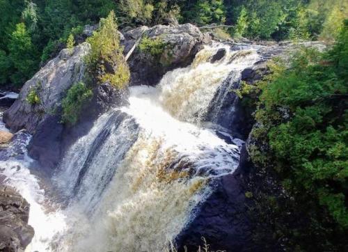 A cascading waterfall flows over rocky cliffs, surrounded by lush green trees and a serene natural landscape.