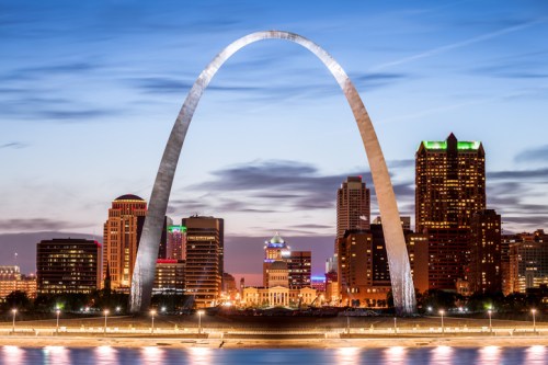 The Gateway Arch illuminated at dusk, with a city skyline in the background and a calm river in the foreground.