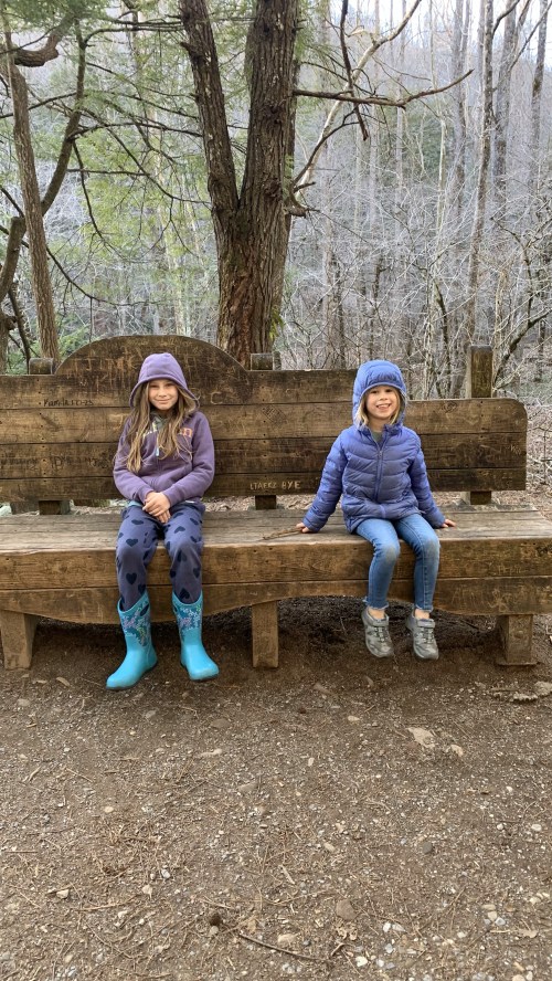 Two children sit on a wooden bench in a forest, wearing winter clothing and boots, surrounded by bare trees.
