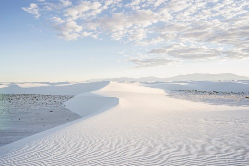A vast white desert landscape with rolling sand dunes under a blue sky and scattered clouds.