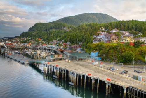 A scenic view of a waterfront town with colorful buildings, a dock, and lush green mountains in the background.