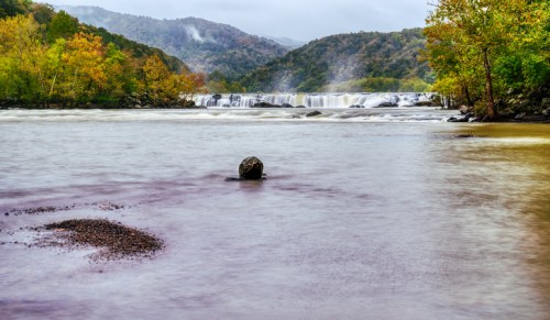 A serene river scene with a waterfall, surrounded by colorful autumn foliage and misty mountains in the background.