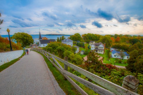 A scenic view of a winding path through a colorful village, with a church steeple and ocean in the background.