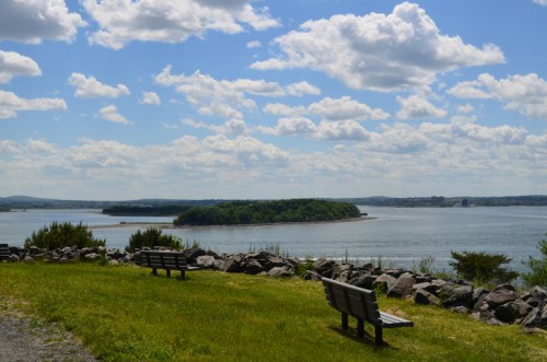 Scenic view of a river with an island, featuring two benches and a grassy area under a partly cloudy sky.
