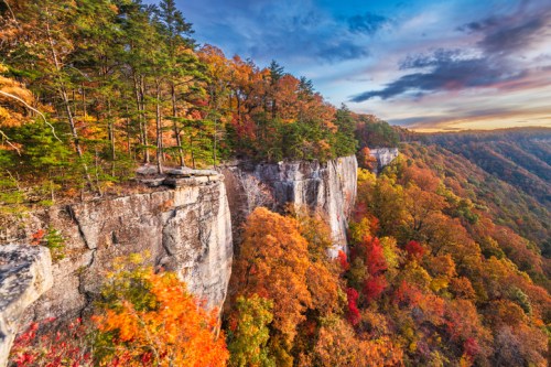 A scenic view of a cliff surrounded by vibrant autumn foliage under a colorful sky at sunset.