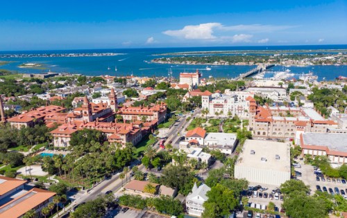 Aerial view of a coastal town with red-roofed buildings, lush greenery, and a marina under a clear blue sky.