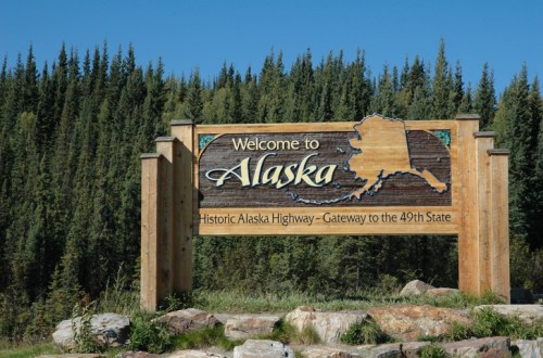 Wooden welcome sign for Alaska, featuring the state outline and text about the Historic Alaska Highway. Surrounded by trees.