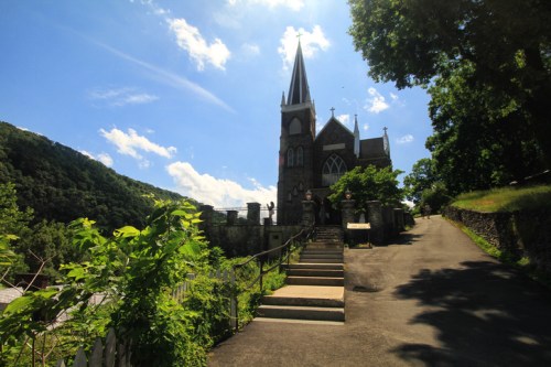 A stone church with a tall spire, surrounded by greenery and hills under a bright blue sky.