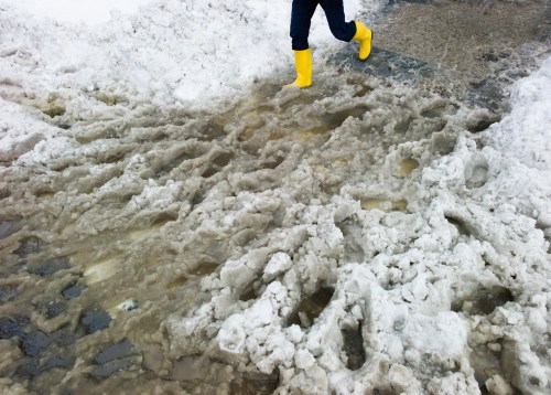 A person in yellow boots walks through slushy snow and muddy water on a winter day.
