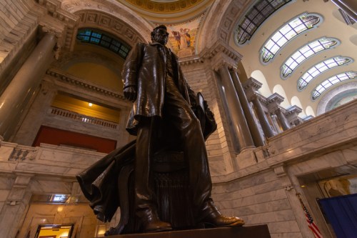 A large bronze statue of a man in a suit stands in a grand, marble-finished hall with ornate architecture.