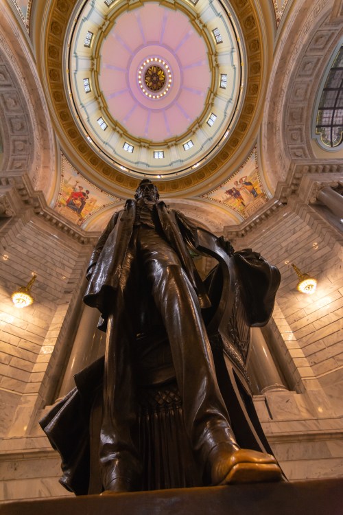 A bronze statue stands tall beneath a beautifully decorated dome with intricate designs and soft lighting.