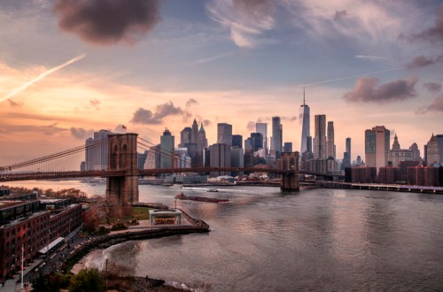A sunset view of the Brooklyn Bridge with the Manhattan skyline in the background, reflecting on the water.