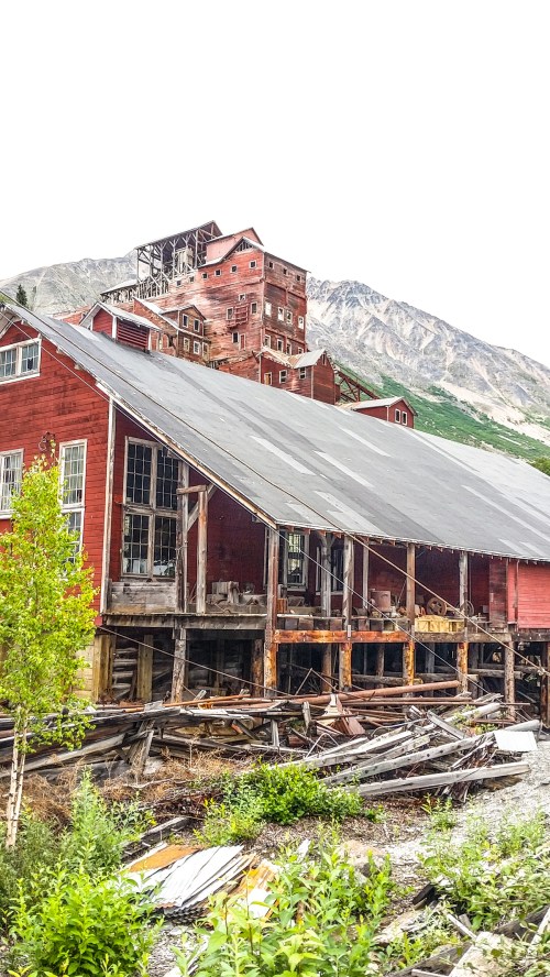 Abandoned red buildings with a mountain backdrop, showcasing weathered wood and overgrown vegetation.