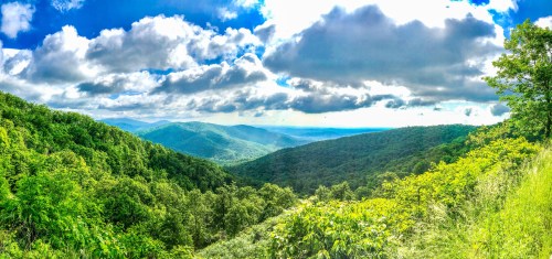 A panoramic view of lush green mountains under a bright blue sky with fluffy clouds.