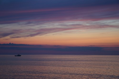 A tranquil seascape at sunset, featuring soft pink and purple clouds reflecting on calm waters with a small boat in the distance.