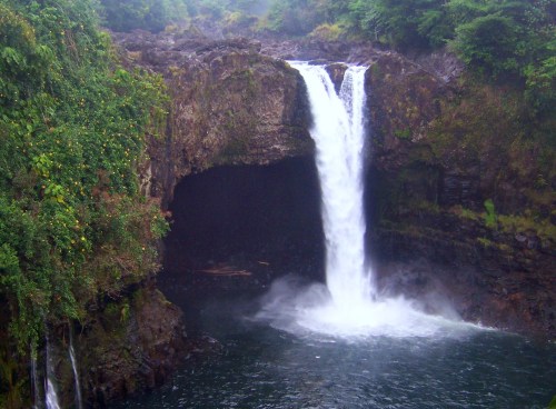 A waterfall cascades into a dark cave, surrounded by lush greenery and misty atmosphere.