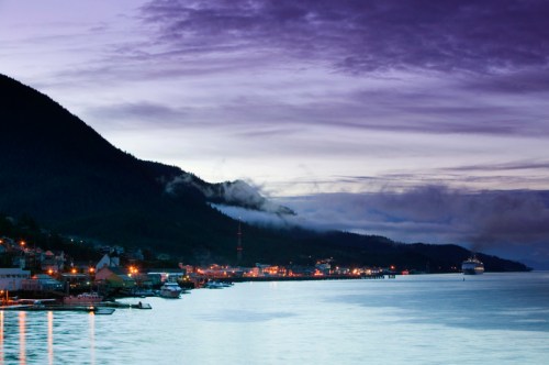 A serene coastal town at dusk, with mountains, clouds, and lights reflecting on the calm water.