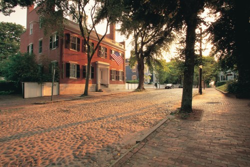 A quiet street lined with trees, featuring a brick building with an American flag and cobblestone pavement at sunset.