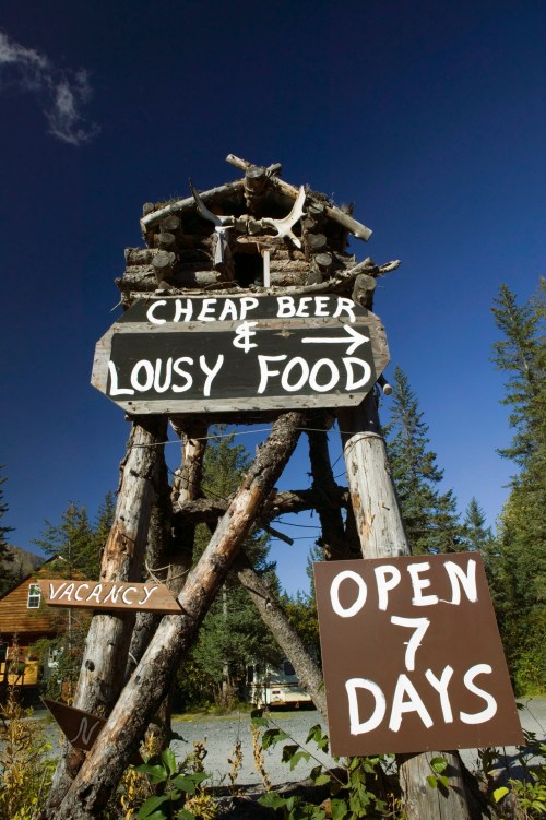 A rustic wooden sign advertising "Cheap Beer & Lousy Food," with arrows and "Open 7 Days" below.