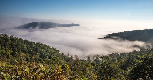 A panoramic view of lush green mountains shrouded in mist, with a clear blue sky above.