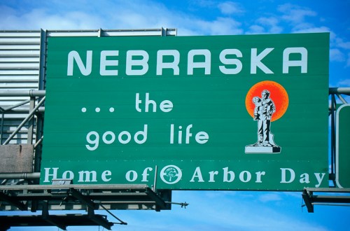 Green highway sign reading "Nebraska... the good life, Home of Arbor Day," featuring a statue and a bright orange sun.