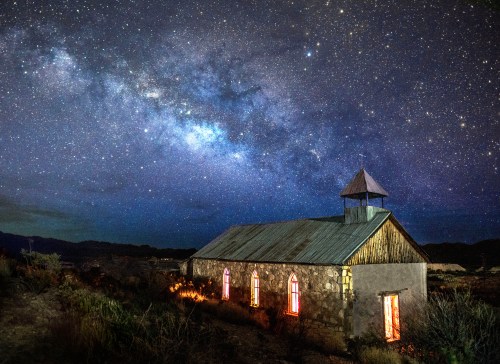 A stone church under a starry night sky, with colorful windows and the Milky Way visible above.