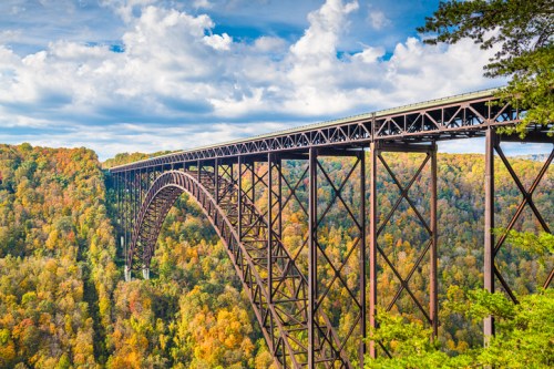 A large metal bridge spans a colorful forested valley under a blue sky with fluffy clouds.