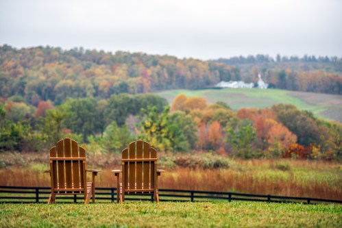 Two wooden Adirondack chairs face a scenic view of rolling hills and autumn foliage under a cloudy sky.