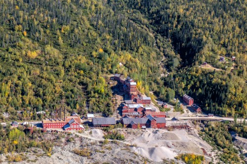 Aerial view of an old mining complex surrounded by lush green trees and hills, with buildings and smokestacks visible.