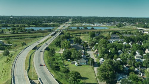 Aerial view of a highway winding through green fields and a small residential area near a body of water.