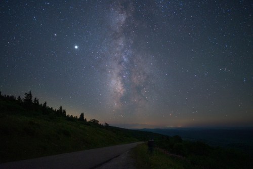 A starry night sky filled with the Milky Way, viewed from a quiet road surrounded by trees and hills.