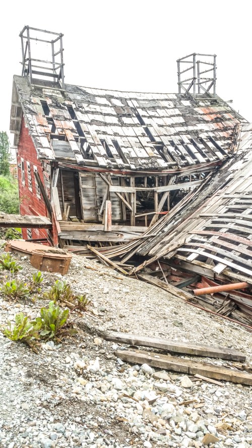 A dilapidated red barn with a sagging roof and exposed wooden beams, surrounded by gravel and overgrown plants.