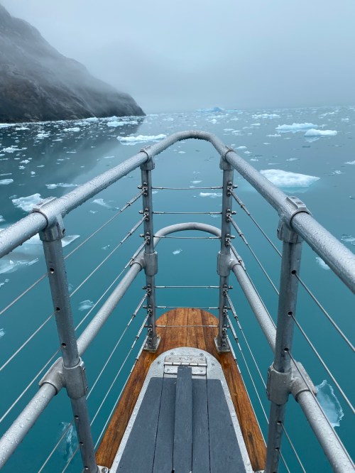 A metal and wooden boat deck extends over icy blue waters, surrounded by floating ice and misty mountains.