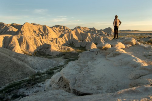 A person stands on a rocky ledge, overlooking rugged, sunlit badlands under a clear sky.