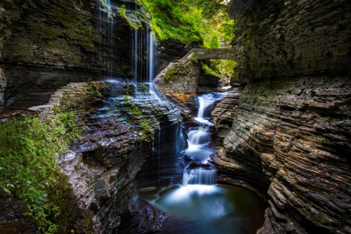 A serene waterfall cascades over rocky cliffs, surrounded by lush greenery and a stone bridge in a tranquil gorge.