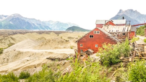Abandoned red building surrounded by mountains and sandy terrain, with greenery in the foreground.