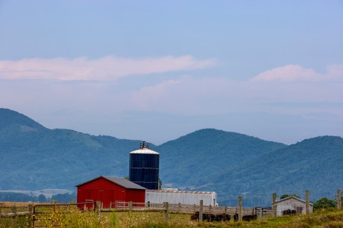 A red barn and silo sit in a rural landscape with rolling green hills under a blue sky.
