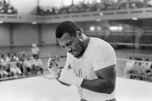 A black and white photo of a boxer in a gym, preparing for a match, with spectators in the background.