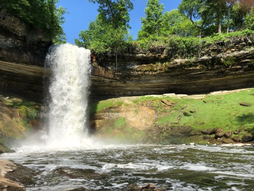 A cascading waterfall flows over rocky cliffs into a serene pool, surrounded by lush greenery and blue skies.